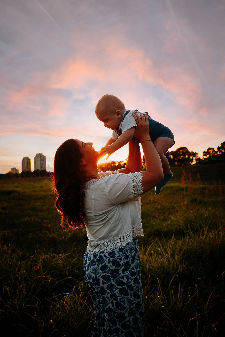 Mlada mama stoji sa sinom u travi u ružičastom zalasku sunca